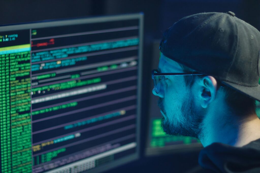 Cybersecurity analyst watching code and system logs on multiple monitors in a dark room.