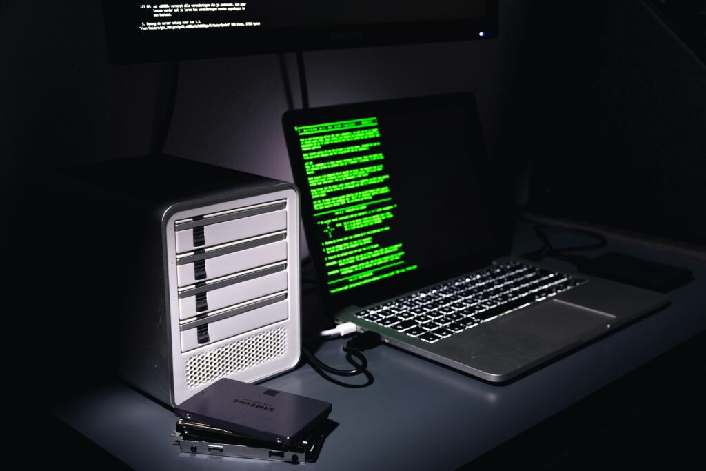 Laptop displaying code next to external storage and hard drives, representing cybersecurity monitoring and backups.