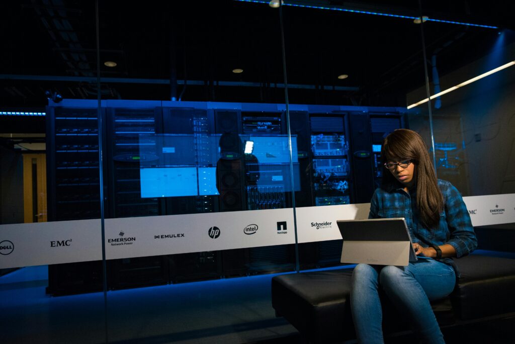 IT professional working on a laptop near server racks while reviewing infrastructure status for NOC services.