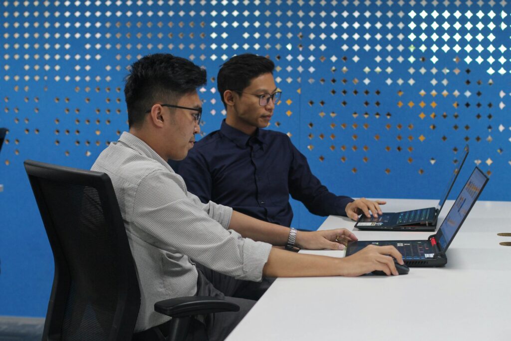 Two coworkers working on laptops at a shared desk in an office.