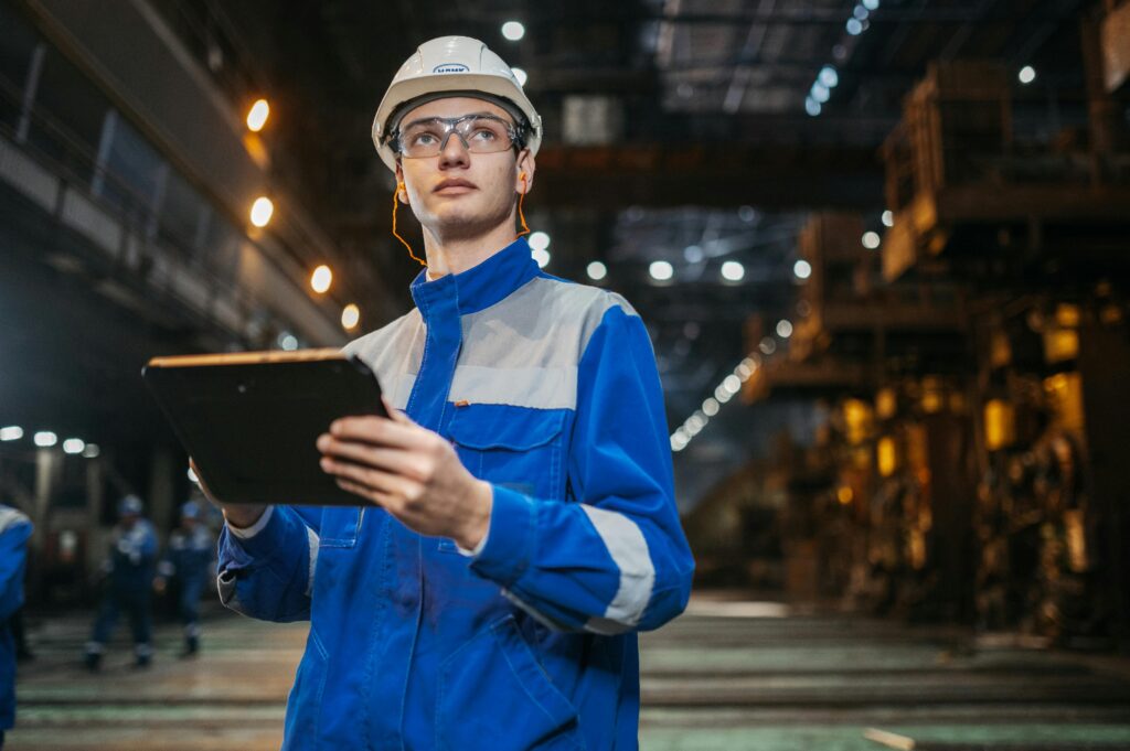 Engineer in safety gear using a tablet on a large industrial manufacturing floor.