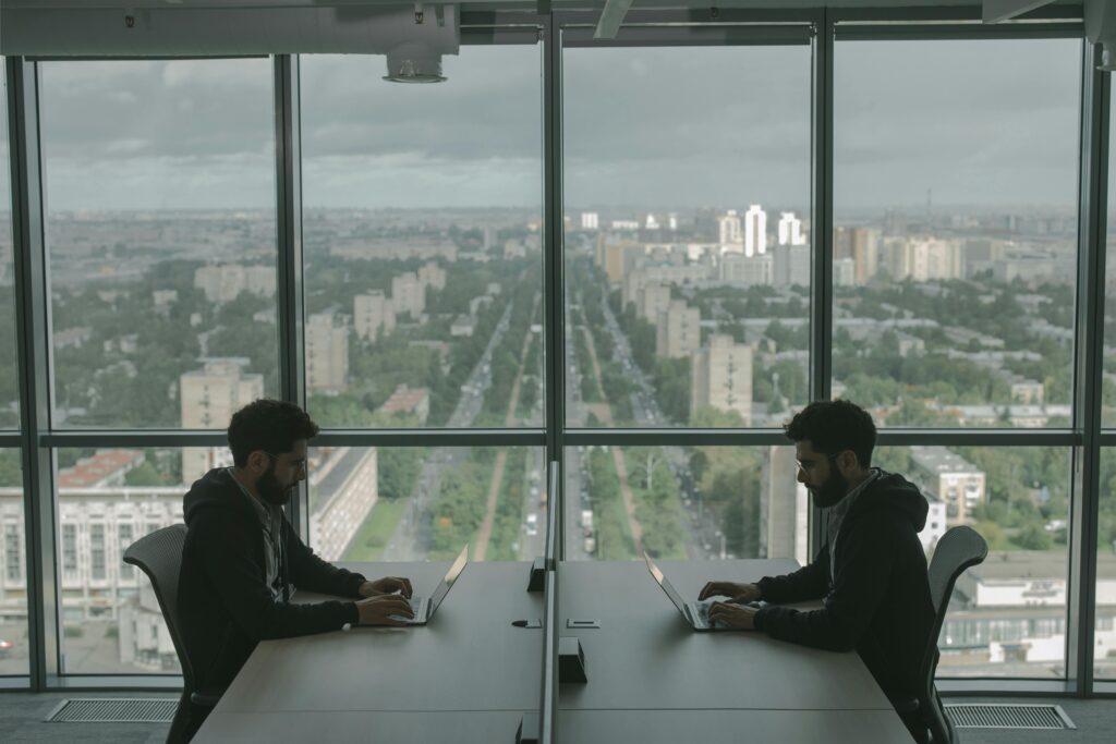Two professionals working on laptops across from each other in a glass office with a large city view
