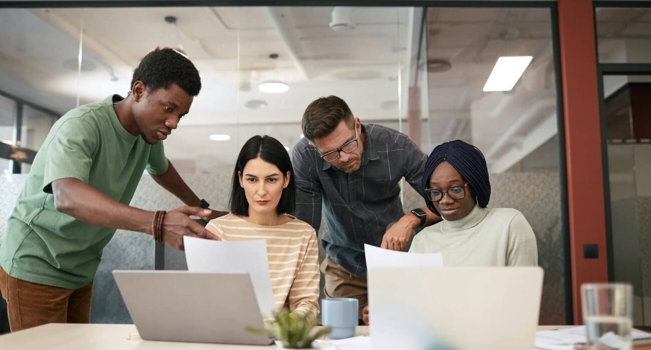 Four employees stand around a table reviewing documents and laptops during a workplace security awareness discussion.