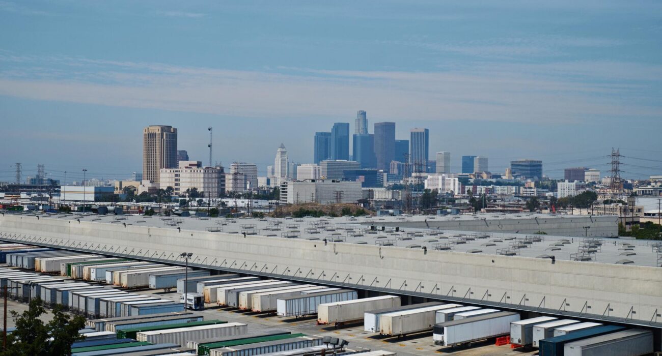 Large warehouse and distribution center with freight trailers and the Downtown Los Angeles skyline in the background.